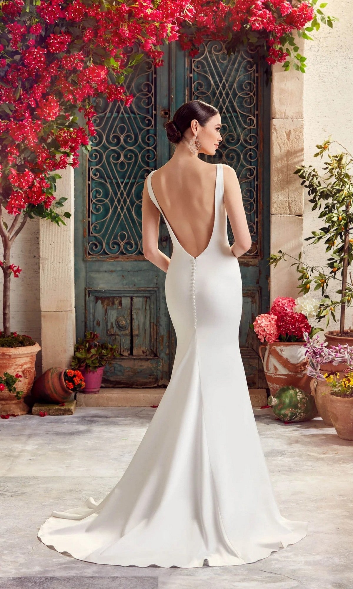 Wearing the Long White Dress: Alyce 7052, a modern bride stands with her back to the camera before an ornate green door, surrounded by pots of vibrant pink flowers.